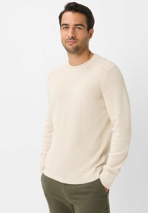 Uomo con capelli scuri che indossa un maglione beige a maniche lunghe e pantaloni verdi, in piedi con le mani in tasca davanti a uno sfondo bianco.