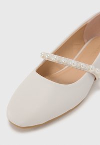 Pantofii plata din piele artificiala alba, cu varf patratos si o curea decorata cu perle. Textura neteda cu un captusala interioara de nuanta bej deschis.