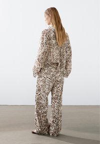 Žena s dlouhými vlasy, oblečená v volné, puntíkované béžové blůze a odpovídajících širokých kalhotách, stojí čelem k jednoduché bílé zdi na šedé podlaze.