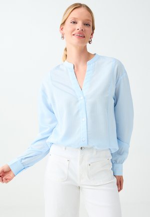 Kobieta ubrana w jasnoniebieską zapinaną na guziki bluzkę i białe spodnie, z srebrnymi kolczykami kołami, stojąca na prostym jasnym tle.
