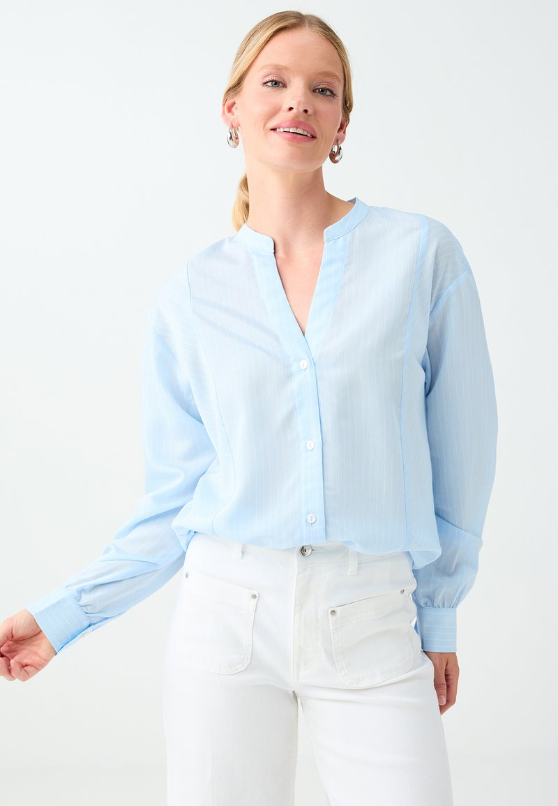 Femme portant un chemisier bleu clair boutonné et un pantalon blanc, avec des boucles d'oreilles créoles argentées, debout devant un fond clair uni.