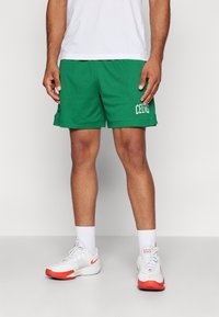 Gröna nätbasketshorts med sidofickor och vit text "CELTICS"; matchas med vita sneakers med röda accenter och vita strumpor.