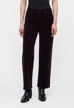 Personne portant un pantalon en velours côtelé violet foncé à jambes droites associé à des bottines noires à talons sur fond blanc uni.