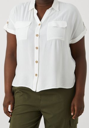 Persona con una camisa blanca de manga corta con botones y dos bolsillos en el pecho y pantalones verde oliva con bolsillos laterales.