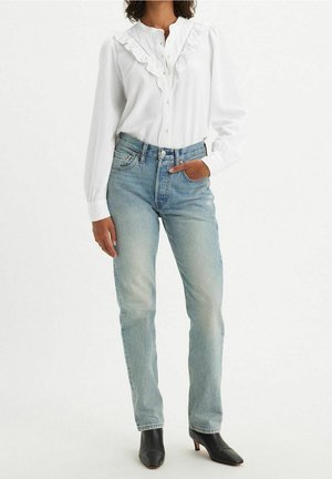 Femme portant un chemisier blanc à volants, un jean taille haute bleu clair et des bottines noires à talons, debout avec une main dans la poche.