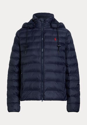 Młoda kobieta nosząca granatową pikowaną kurtkę, czarny sweter, luźne białe spodnie i białe trampki, stoi z rękami w kieszeniach.