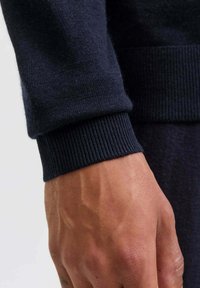 Primo piano della mano e del polso di una persona che indossa un maglione a maglia di un blu navy scuro, con polsini e vita a costine.