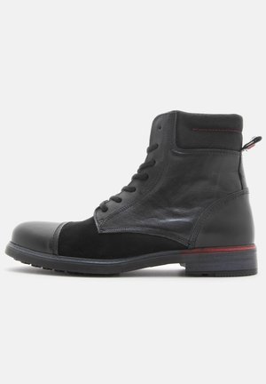 Black leather and suede ankle boot with lace-up front, padded collar, pull tab, and red stitching detail on heel and sole edge.