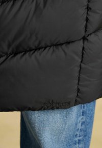 Czarna kurtka puchowa o pikowanej fakturze z haftowanym logo "JOULES". Noszona na niebieskich dżinsach z kontrastowym przeszyciem.
