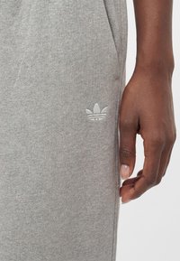Gros plan d'une main à la peau foncée à côté de la poche d'un pantalon de survêtement gris clair avec un petit logo blanc Adidas.
