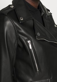 Gros plan sur une veste en cuir noir de motard avec une poche zippée argentée et des boutons-pression, portée par-dessus un col roulé noir.