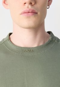 Olíva zöld póló, kerek nyakkivágással és hímzett "BOSS" logóval a galléron. Az anyag simának és testhezállónak tűnik.