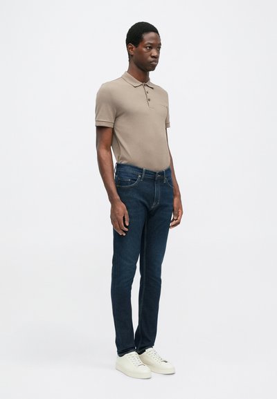 Beige pólóing, három gombbal, sötétkék slim-fit farmernadrággal és fehér sportcipővel párosítva. Egyszerű dizájn, sokoldalú szín és sima textúra.