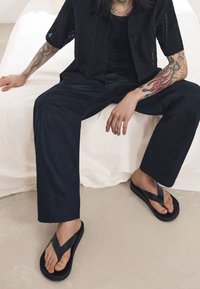 Sandale negre cu talpă groasă și curele în formă de V, asortate cu haine negre, așezate pe o suprafață de culoare neutră.