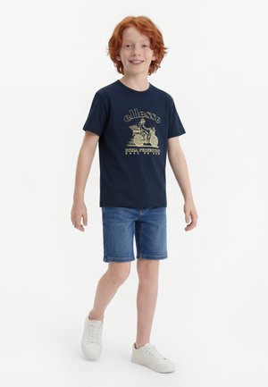 Αγόρι με κόκκινα μαλλιά που περπατά φορώντας μπλε σκούρο γραφικό μπλουζάκι, μπλε τζιν σορτς και λευκά αθλητικά παπούτσια σε απλό λευκό φόντο.