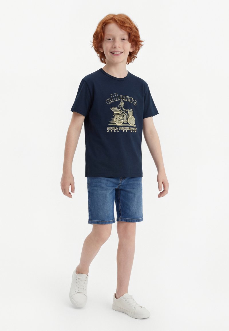 Αγόρι με κόκκινα μαλλιά που περπατά φορώντας μπλε σκούρο γραφικό μπλουζάκι, μπλε τζιν σορτς και λευκά αθλητικά παπούτσια σε απλό λευκό φόντο.