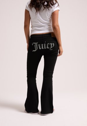Woman wearing black flared jeans with "Juicy" in rhinestones on the back pockets and a white short-sleeve shirt, standing against light background.