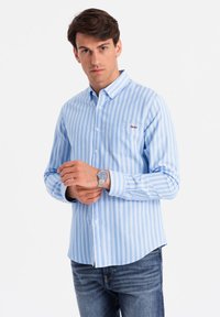 Camisa de botones a rayas azul claro y blanca, con un bolsillo en el pecho, corte casual y puños de botón, combinada con jeans oscuros y un reloj de plata.