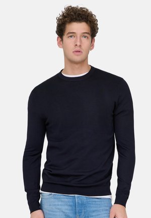 Ung mand med krøllet hår iført en sort rundhalset sweater over en hvid skjorte og lyseblå jeans, stående mod en ensfarvet baggrund.
