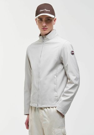 Giovane uomo con giacca leggera grigia con cerniera e pantaloni beige, indossa un berretto da baseball marrone con la scritta "Colmar Originals".