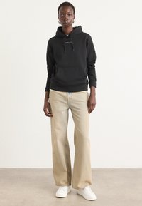 Sudadera negra con capucha y bolsillo canguro, combinada con jeans de pierna ancha en un tono beige claro y zapatillas blancas, contra un fondo neutro.