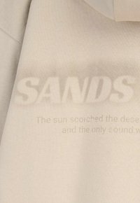 Beżowa tkanina z dużym, wyblakłym napisem "SAVDS" i mniejszym, częściowo widocznym tekstem poniżej, który brzmi "Słońce przypaliło pustynię..."