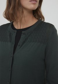 Tamnozeleni kardigan s dugmadi, s teksturiranim pletenim dizajnom na ramenima, rebrastim manžetama i glatkim materijalom.