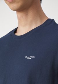 Tummansininen puuvillapaita, jossa on pyöreä kaula-aukko ja lyhyet hihat. Paidassa on pieni valkoinen logo, jossa lukee "Marc O'Polo DENIM."
