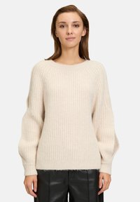 Maglione di colore crema lavorato a maglia con texture a coste, scollatura rotonda e maniche ampie e leggermente voluminosi. Abbinato a pantaloni neri.