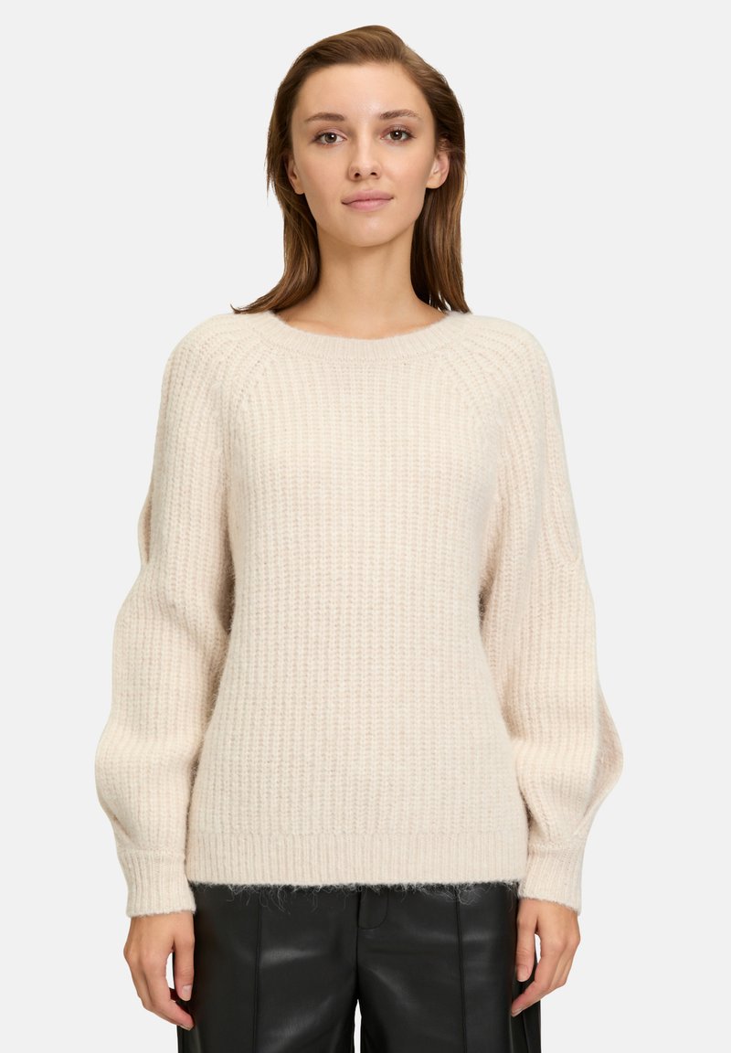 Maglione di colore crema lavorato a maglia con texture a coste, scollatura rotonda e maniche ampie e leggermente voluminosi. Abbinato a pantaloni neri.