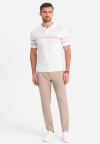 Polo blanc à manches courtes avec des rayures beiges, associé à un pantalon à carreaux beige. Texture lisse, coupe ajustée et baskets blanches complètent le look.