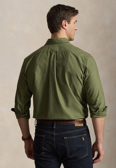 Muž v olivovo zelenej košeli s vyhrnutými rukávmi a tmavomodrých džínsoch, stojaci chrbtom k neutrálnemu pozadiu.