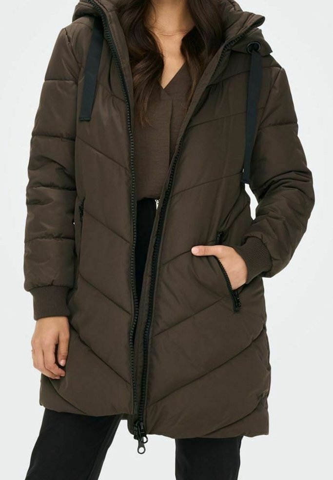Vrouw in een lange, donkerbruine gewatteerde winterjas met een capuchon, rits aan de voorkant, met rits gesloten zijzakken en geribbelde manchetten, hand in de zak.