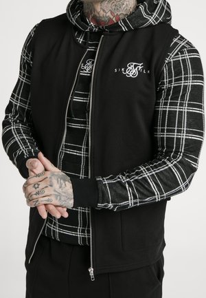 Zwarte ritssluitingvest over een zwart-witte geruite lange mouwen shirt. Heeft een capuchon, geribbelde manchetten en logo detail op het vest.