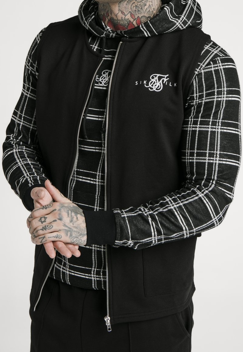 Veste sans manches noire zippée avec capuche, portée sur une chemise à carreaux noir et blanc à manches longues. Présente un logo sur la poitrine et des mains tatouées jointes devant.