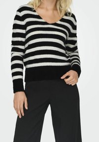 Pull duveteux rayé noir et blanc avec un col en V, manches longues et coupe décontractée, associé à un pantalon noir.