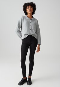 Cardigan gris à côtes avec fermeture à boutons et poche, associé à un jean noir ajusté et des chaussures à lacets noires. Texture douce, design décontracté.