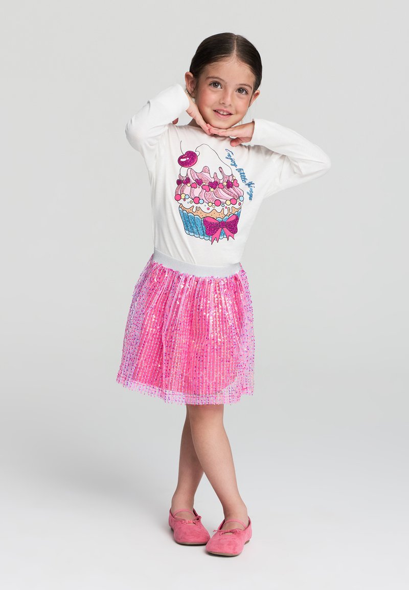 Biała koszula z długim rękawem z grafiką babeczki, zestawiona z różową spódnicą wyszywaną cekinami. Różowe miękkie buty dopełniają strój.