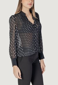 Blusa nera trasparente con pois bianchi, caratterizzata da un design plissettato, colletto e chiusura con bottoni. Maniche lunghe con polsini aderenti.