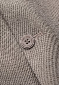 Gros plan d'un bouton en tissu gris cousu sur un tissu tissé marron clair texturé avec le mot « OMBRE » gravé à plusieurs reprises sur le bouton.