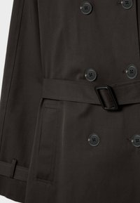 Część ciemno-brązowego płaszcza trench, pokazująca podwójne zapięcia na guziki, pasek z prostokątną klamrą oraz boczną kieszeń z klapką.