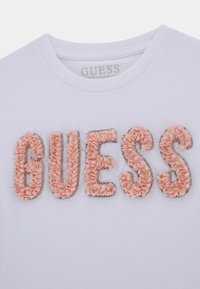 Vit ekologisk bomulls t-shirt med "GUESS" i dimensional rosa tyg med silverpärlor som accent, rund halsringning och korta ärmar.