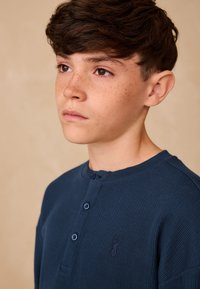 Tumši zils ribots krekls ar garām piedurknēm, trīs pogām pie kakla un izšūtu logotipa detaļu. Gluda tekstūra un klasiskā piegrieztne.