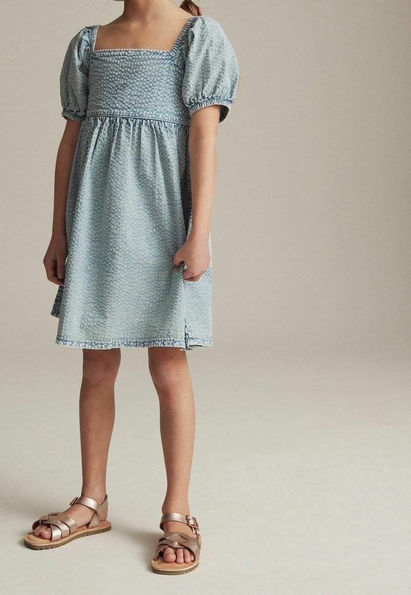 Enfant portant une robe bleu clair texturée avec des manches bouffantes et un décolleté carré, associée à des sandales métalliques or rose, debout sur un sol neutre.