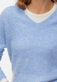 Pull tricot bleu clair doux à col en V porté sur un t-shirt blanc à col rond sur une personne aux cheveux châtain clair.