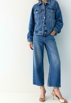 Persoon gekleed in een blauwe spijkerjas en wijd uitlopende jeans met gouden sandalen met hoge hakken, staand tegen een eenvoudige lichte achtergrond.