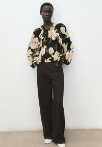 Blusa nera floreale con maniche a sbuffo, pantaloni larghi marroni e scarpe color crema. L'outfit presenta grandi fiori rosa chiaro e foglie verdi.