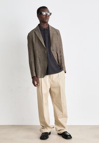 Sacou dintr-un material texturat de culoare brună, polo negru, pantaloni bej și pantofi negri masivi. Model purtând ochelari de soare cu un design unghiular. Poziție în picioare.