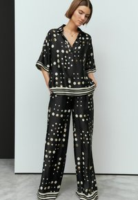 Schwarzes Satin-Outfit mit einem locker sitzenden, kurzärmligen Shirt und weit geschnittenen Hosen, die mit großen beigen und goldenen Punkten sowie gestreiften Akzenten verziert sind.