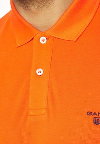 Detailní záběr na jasně oranžové polo tričko se dvěma bílými knoflíky a částí vyšívaného loga v námořnické modři na hrudi.